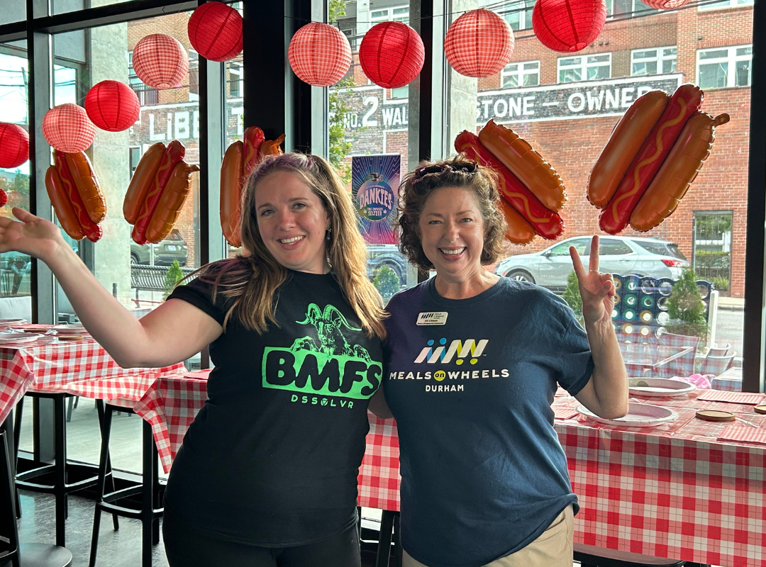 Ansley (left) and Jill Ullman (right) post during Four Feet to Hell Fundraiser by throwing up the piece sign in front of handing hot dog banner.
