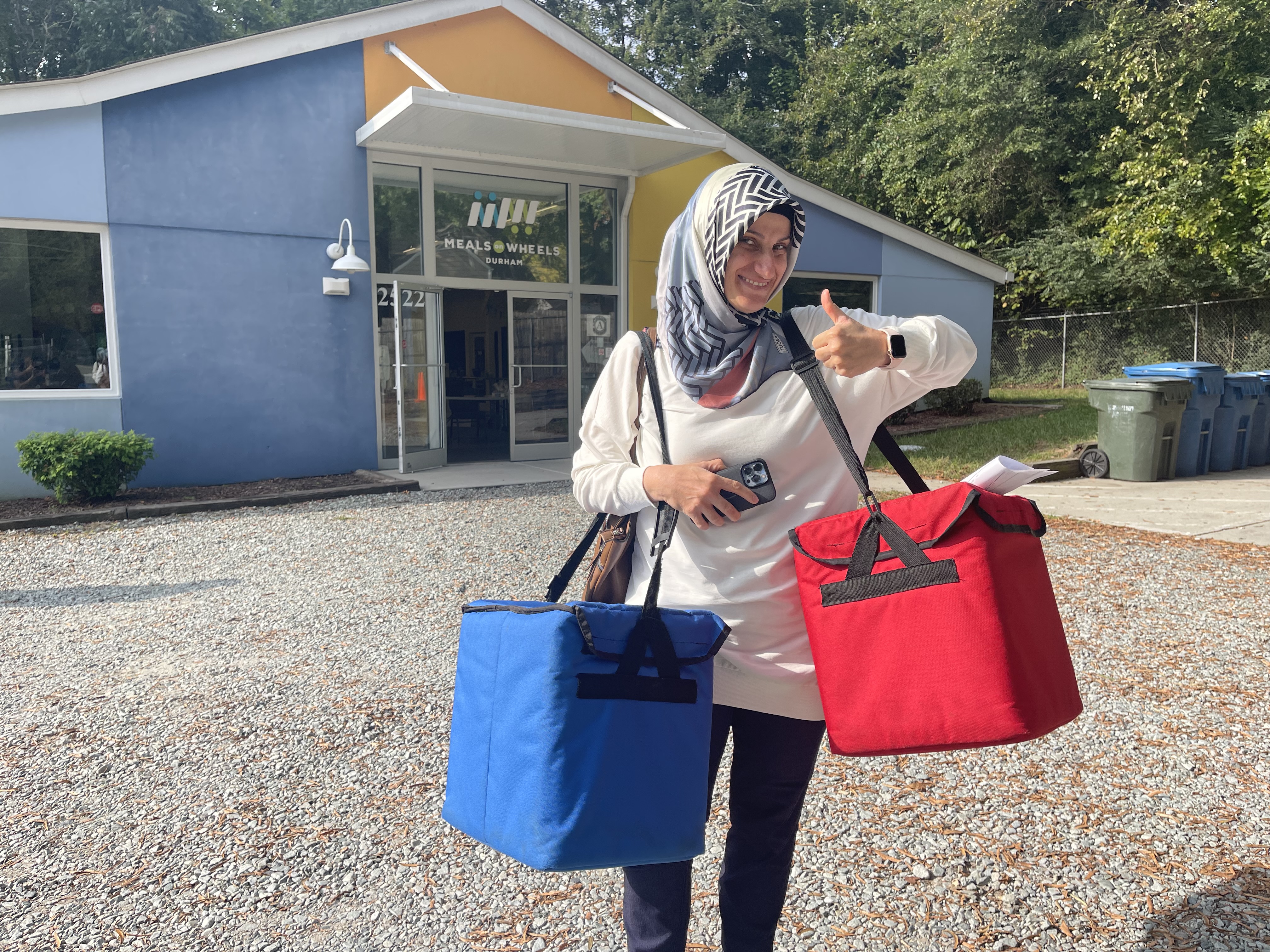 Volunteer smiling with a thumbs up while holding the red and blue meal delivery insulated bags in front of the Meals on Wheels Durham building.